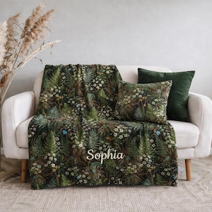 May include: A botanical-themed throw blanket and cushion set with a dark green and brown floral pattern. The blanket is draped over a white sofa, with a matching cushion and a solid green cushion. The name "Sophia" is embroidered on the blanket.