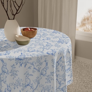 May include: A white round tablecloth with a blue bunny and floral pattern. The tablecloth is on a round table with a bowl of fruit and a vase with a branch.