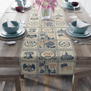 May include: A beige table runner with a repeating pattern of patriotic symbols, including the Statue of Liberty, the Liberty Bell, and the American flag. The text "USA 1776 2026" and "250 2026" are also featured. The runner is placed on a wooden table.