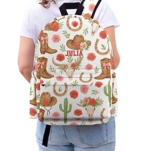 Puede incluir: Mochila blanca con un estampado repetido de botas de vaquero, sombreros, herraduras, rosas y una calavera de toro. El nombre "JULIA" está impreso en rojo. La mochila tiene correas negras y un bolsillo delantero blanco.