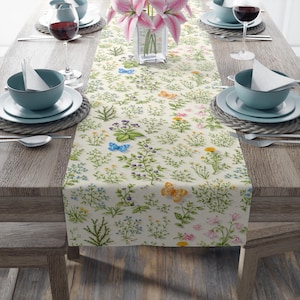 May include: A floral table runner with a cream background, featuring a pattern of wildflowers and butterflies in shades of blue, yellow, and orange. The runner is set on a wooden table with place settings, wine glasses, and a vase of pink lilies.