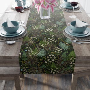 May include: A dining table set for a meal, featuring a floral table runner with a dark background and botanical design. The runner showcases ferns, flowers, and other foliage. The table is set with plates, bowls, silverware, and wine glasses. A vase of pink lilies is in the centre.