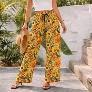 May include: Yellow floral print palazzo pants with a black drawstring. The pants feature a repeating pattern of sunflowers and other flowers in shades of yellow, orange, and green. A straw hat is held in the left hand.