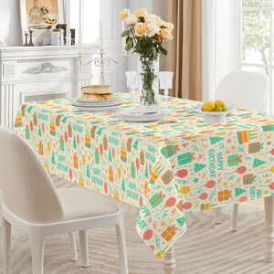 May include: A rectangular tablecloth with a festive birthday theme. The cream-colored fabric is covered with colorful illustrations of gifts, balloons, party hats, and cakes. The words "Happy Birthday" are printed throughout the design.