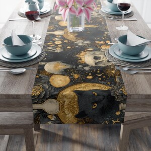 May include: A table runner with a dark, celestial design featuring a black cat, skulls, and gold accents. The runner is set on a wooden table with teal tableware and wine glasses. A vase of pink lilies adds a floral touch.