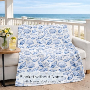 Puede incluir: Una manta blanca con un diseño de conchas marinas azules, colocada sobre un sofá blanco en un balcón con vistas al mar. La manta presenta ilustraciones de conchas y el texto "Blanket without Name with Name label available".