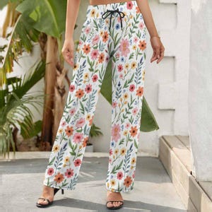 May include: White palazzo pants with a colorful floral pattern. The pants feature a drawstring waist and a vibrant design of orange, pink, yellow, and blue flowers with green leaves. The pants are made of a lightweight fabric.
