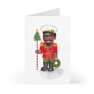 May include: Watercolor Christmas card featuring a nutcracker soldier in a red uniform with gold accents, a white hat, and a black mustache. The nutcracker holds a Christmas tree and a wreath. The card is on a white background.