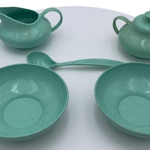 May include: A set of four teal green speckled melamine bowls and a matching creamer and sugar bowl. The set includes two large bowls, a smaller creamer, and a sugar bowl with a lid.