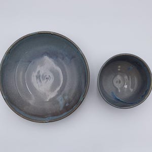 May include: Two handmade ceramic bowls. The larger bowl is a shallow dish with a dark blue and gray glaze, and a brown rim. The smaller bowl is a rounded shape with a similar glaze. Both bowls are on a white background.