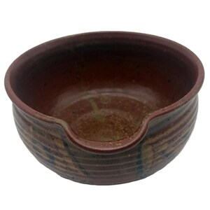 May include: A round, dark brown ceramic bowl with a textured, ribbed exterior and a smooth interior. The rim features a distinct U-shaped notch or spout on one side, and the surface displays subtle variations in color and a speckled finish.