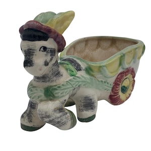 Puede incluir: Un macetero de cerámica con forma de burro tirando de un carro. El burro está pintado de blanco con rayas negras y tiene una corona verde alrededor del cuello. El carro está pintado de verde, amarillo y rojo con un diseño floral.