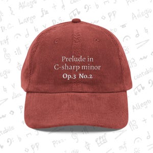 May include: A brick-red corduroy baseball cap. The cap features the text "Prelude in C-sharp minor Op.3 No.2" embroidered in white. The background has musical notes and symbols.