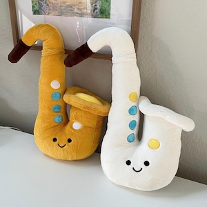 Puede incluir: Dos juguetes de peluche con forma de saxofón. Uno es amarillo con detalles marrones y azules, el otro es blanco con detalles marrones y azules. Ambos tienen caras sonrientes y están sobre una superficie blanca.