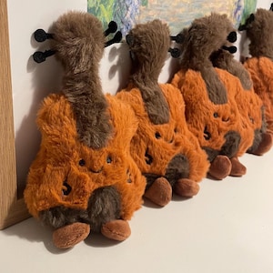 Può includere: Una fila di violini giocattolo di peluche. Ogni giocattolo ha un manico e una paletta marroni, un corpo arancione e piedini marroni. Il corpo ha un numero 5 cucito. I giocattoli sono allineati su una superficie bianca.