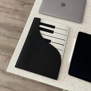 Puede incluir: Una funda negra y blanca con diseño de teclas de piano para una tableta o portátil. La funda tiene un bolsillo negro en la parte delantera y un diseño de teclas de piano en la parte trasera. Un portátil y una tableta están al lado de la funda.