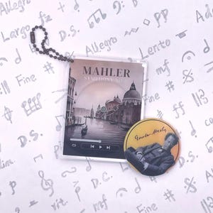 May include: A clear acrylic keychain featuring a cover design for Mahler's Symphony No. 5, with a Venetian canal scene. A round pin with a portrait of Gustav Mahler is also present. The background is decorated with musical notes.