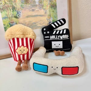 May include: A plush set of movie-themed toys. Includes a popcorn container with red and white stripes, a black clapperboard with "HOLLYWOOF" text, and 3D glasses with blue and red lenses. All toys have smiling faces.