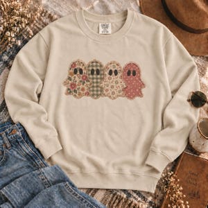 Op de afbeelding: Een beige sweater met ronde hals en een afbeelding van vier geesten op een rij. Elke geest heeft een eigen patroon: bloemen, groen-witte vichy-ruit, kleine bloemetjes en een effen oudroze kleur met witte spikkels.