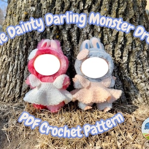 May include: Two plush monster dolls, one pink and one blue, each with a white circle face and wearing a crochet dress. The text "The Dainty Darling Monster Dress" and "PDF Crochet Pattern" are displayed above and below the dolls.