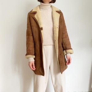 May include: A brown suede coat with a cream-colored shearling lining and trim. The coat features a button closure and a classic lapel collar. The sleeves have shearling cuffs. The coat is worn over a cream-colored turtleneck sweater and cream-colored pants.