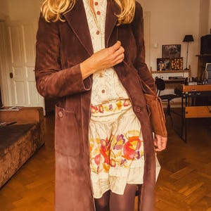 Könnte beinhalten: Ein langer, dunkelbrauner Wildledermantel mit Reverskragen und Knopfverschluss. Der Mantel wird über einer cremefarbenen Bluse und einem Rock mit floralem Muster getragen. Das Outfit wird durch dunkle Strumpfhosen ergänzt.