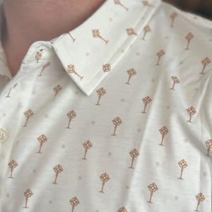 May include: Cream polo shirt with a repeating pattern of brown geometric shapes and small grey dots. The shirt has a button-down collar and short sleeves. The fabric appears to be a lightweight, breathable material.