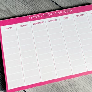 A pink weekly planner notepad with white text. The top of the notepad says 'Things To Do This Week.'  Each day of the week is listed across the top: Monday, Tuesday, Wednesday, Thursday, Friday, Saturday, Sunday.  Rows of blank spaces are provided under each day for writing tasks or appointments. The notepad is a useful tool for organizing and scheduling weekly activities.