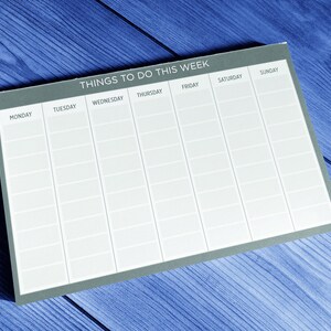 A gray weekly planner on a blue wooden surface. The planner has spaces to write tasks for each day of the week: Monday, Tuesday, Wednesday, Thursday, Friday, Saturday, and Sunday.  The top of the planner says 'Things To Do This Week'.