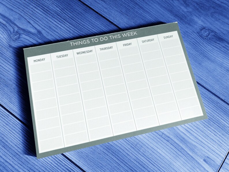 A gray weekly planner on a blue wooden surface. The planner has spaces to write tasks for each day of the week: Monday, Tuesday, Wednesday, Thursday, Friday, Saturday, and Sunday.  The top of the planner says 'Things To Do This Week'.