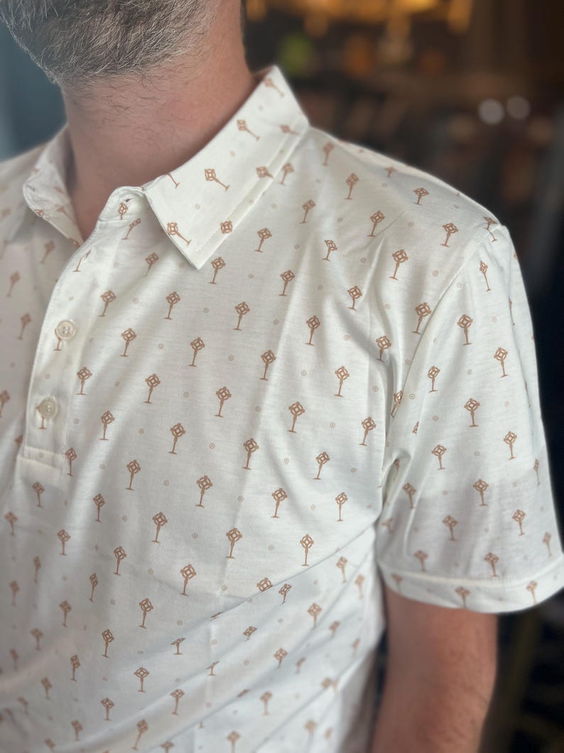 Op de afbeelding: Cr&egrave;mekleurige poloshirt met een herhalend patroon van bruine geometrische vormen en kleine grijze stippen. De shirt heeft een button-down kraag en korte mouwen. De stof lijkt een lichtgewicht, ademend materiaal te zijn.