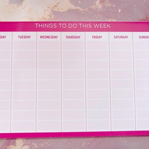 A pink weekly planner with the title 'Things To Do This Week'.  Days of the week are listed across the top: Monday, Tuesday, Wednesday, Thursday, Friday, Saturday, Sunday. Each day has multiple blank lines for writing tasks or appointments. The planner is rectangular and features a simple, clean design.