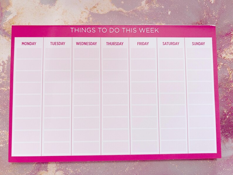 A pink weekly planner with the title 'Things To Do This Week'.  Days of the week are listed across the top: Monday, Tuesday, Wednesday, Thursday, Friday, Saturday, Sunday. Each day has multiple blank lines for writing tasks or appointments. The planner is rectangular and features a simple, clean design.