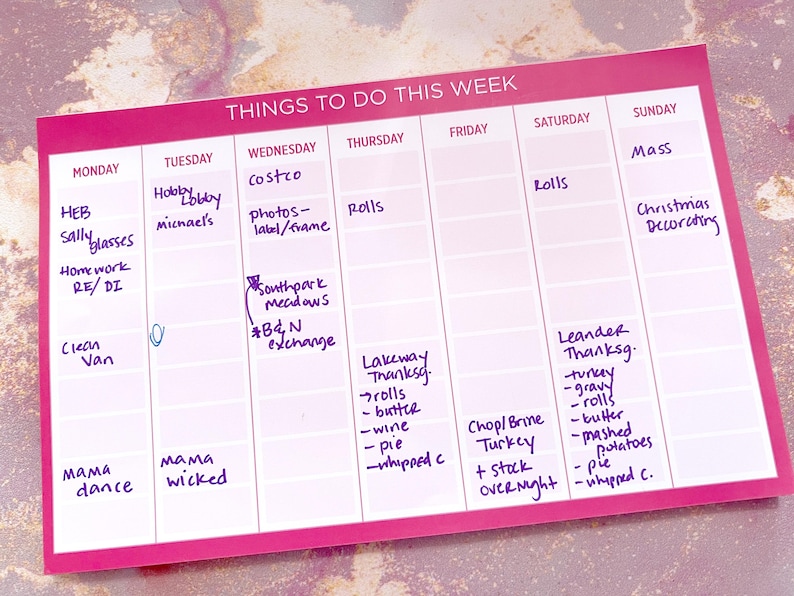 A pink weekly planner with daily sections for scheduling tasks.  The planner includes handwritten entries for various errands, appointments, and Thanksgiving preparations, such as grocery shopping, cooking, and decorating.  The text 'Things To Do This Week' is at the top.  Days of the week are listed across the top, from Monday to Sunday.  Specific tasks include: HEB, Sally glasses, Hobby Lobby, Michael's, Costco, photos-label/frame, rolls, Mass, Christmas decorating, Thanksgiving, and more.