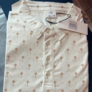 Op de afbeelding: Een cr&egrave;mekleurige poloshirt met een herhaald patroon van bruine, gestileerde sleutels. De shirt heeft een button-down kraag en een label met "THE DAILY Refrain". De shirt is netjes opgevouwen.