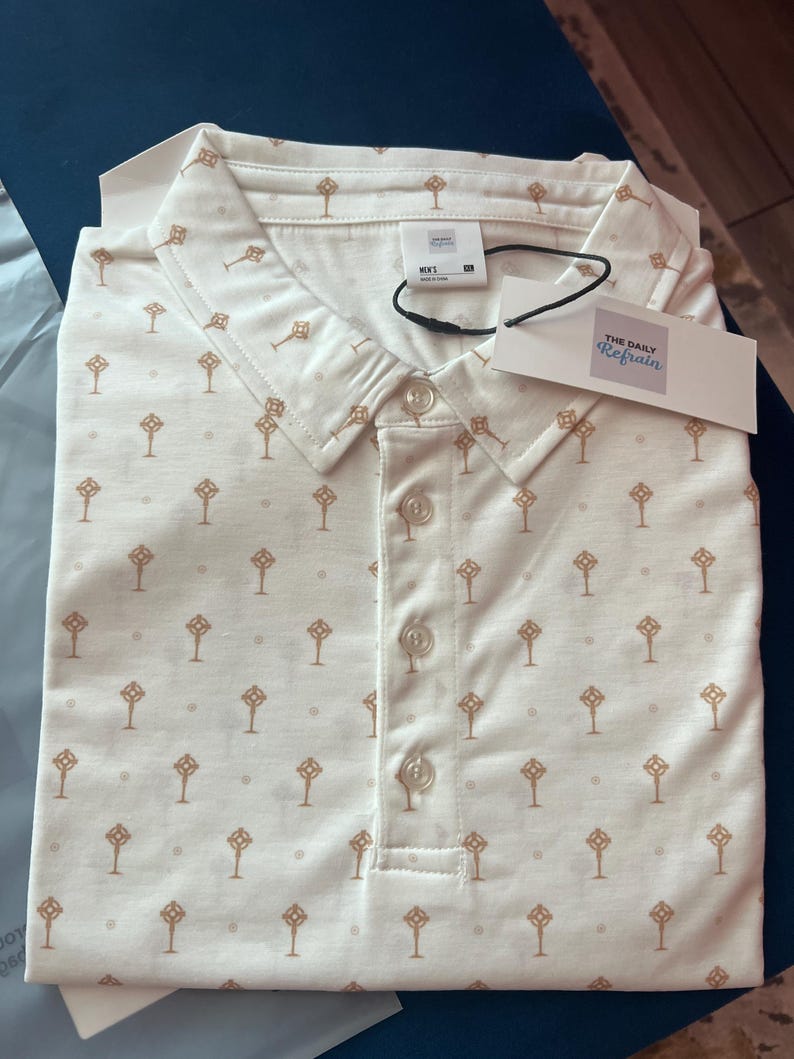 Op de afbeelding: Een cr&egrave;mekleurige poloshirt met een herhaald patroon van bruine, gestileerde sleutels. De shirt heeft een button-down kraag en een label met "THE DAILY Refrain". De shirt is netjes opgevouwen.