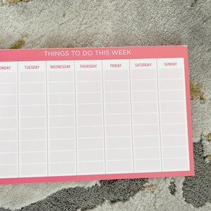 A pink weekly planner with the title 'Things To Do This Week'.  The planner is divided into seven columns for each day of the week: Monday, Tuesday, Wednesday, Thursday, Friday, Saturday, and Sunday. Each day has multiple rows for writing tasks or appointments. The planner is blank and ready to be filled in.