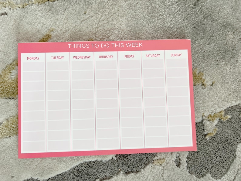 A pink weekly planner with the title 'Things To Do This Week'.  The planner is divided into seven columns for each day of the week: Monday, Tuesday, Wednesday, Thursday, Friday, Saturday, and Sunday. Each day has multiple rows for writing tasks or appointments. The planner is blank and ready to be filled in.