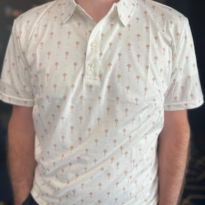 Op de afbeelding: Een witte poloshirt met een herhaald patroon van kleine, bruine boomachtige vormen. De shirt heeft korte mouwen, een kraag en een knoopsluiting met drie knopen. De persoon draagt een marineblauwe broek.