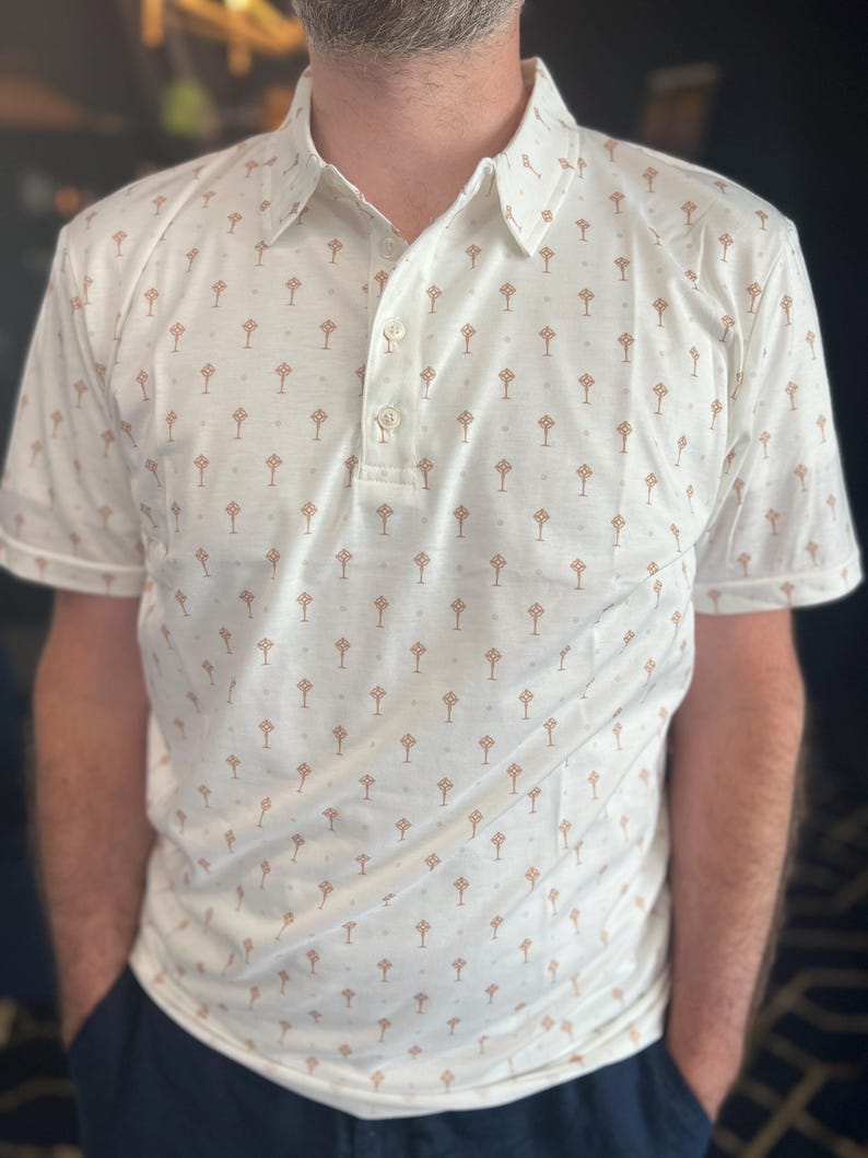 Op de afbeelding: Een witte poloshirt met een herhaald patroon van kleine, bruine boomachtige vormen. De shirt heeft korte mouwen, een kraag en een knoopsluiting met drie knopen. De persoon draagt een marineblauwe broek.