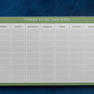A weekly planner notepad with a sage green border. The days of the week are listed across the top: Monday, Tuesday, Wednesday, Thursday, Friday, Saturday, Sunday.  Each day has multiple blank lines for writing tasks or appointments. The title at the top reads: 'Things To Do This Week'.