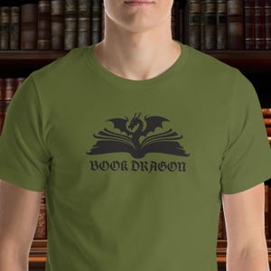 Op de afbeelding: Olijfgroen T-shirt met een zwarte afbeelding van een draak boven een open boek, met de tekst "BOOK DRAGON" eronder. Het ontwerp is gecentreerd. Het shirt heeft een ronde hals en korte mouwen.