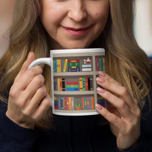 Pode incluir: Caneca de cerâmica branca com uma ilustração colorida de uma estante cheia de livros. A estante tem três prateleiras e está cheia de livros de várias cores. Também há alguns itens decorativos nas prateleiras, como um relógio de areia, uma garrafa e uma planta.