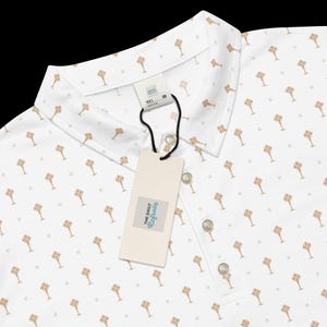 Op de afbeelding: Witte poloshirt met een herhalend goudkleurig sleutelpatroon en kleine stippen. De blouse heeft een kraag, drie knopen en een label met "The Daily Refrain".