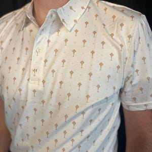 Op de afbeelding: Cr&egrave;mekleurige poloshirt met een herhalend patroon van bruine geometrische vormen. De shirt heeft een button-down kraag en korte mouwen. De stof lijkt een lichtgewicht materiaal te zijn.