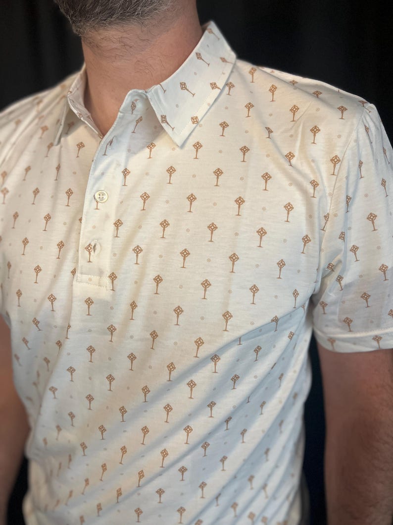 Op de afbeelding: Cr&egrave;mekleurige poloshirt met een herhalend patroon van bruine geometrische vormen. De shirt heeft een button-down kraag en korte mouwen. De stof lijkt een lichtgewicht materiaal te zijn.