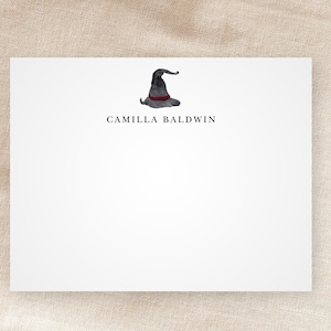 May include: A white card with a black and red witch's hat illustration and the text "CAMILLA BALDWIN" printed in black.