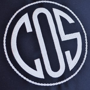 May include: A navy blue fabric with a white embroidered monogram of the letters "COS" in a circle.
