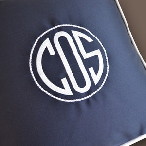 May include: A navy blue pillow with a white monogram of the letters "COS" in a circle. The pillow has a white piping around the edge.