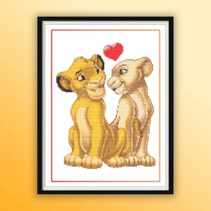 May include: A framed cross-stitch picture showing two lion cubs, one golden yellow and the other beige, with a red heart above them. The cubs are close together, suggesting affection. The artwork is framed in a black frame with a white border.